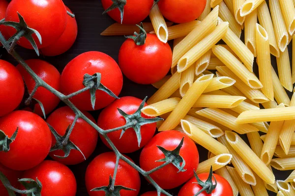 Kleine rode tomaten op de wijnstok, droge penne pasta, op zwart hout f — Stockfoto