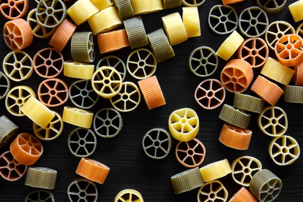 Dry coloured wagon wheel pasta on black textured wood, from abov — ストック写真