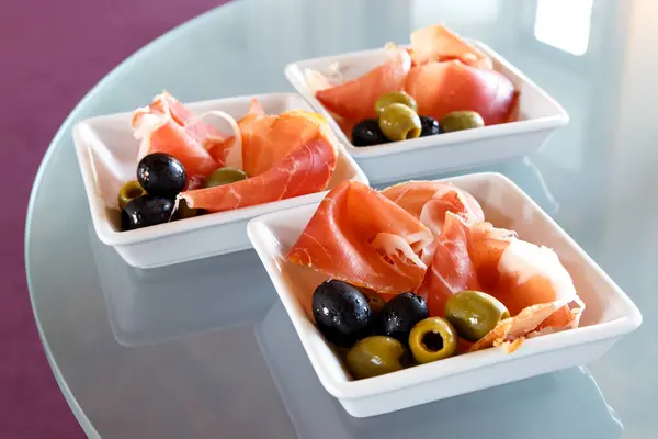 Prosciutto parma ham with olives in small dishes on glass table — Stock Photo, Image