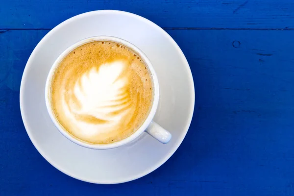 Cappuccino v bílém keramickém šálku s podšálkem na modrém lakovaném dřevě — Stock fotografie