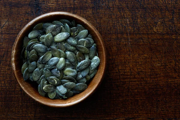 Graines de citrouille séchées pelées dans un bol en bois foncé isolé sur fond foncé — Photo