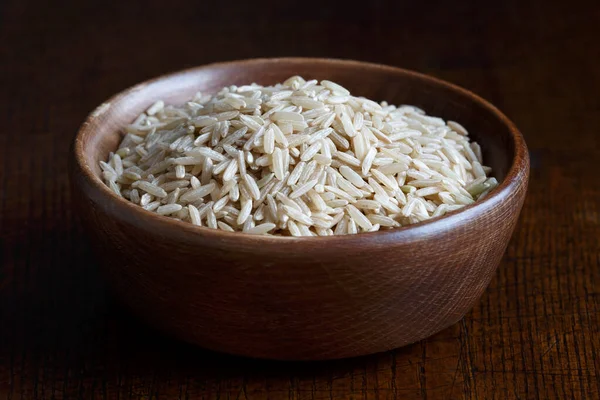 Dry long grain brown rice in brown wooden bowl isolated on dark — Stock Photo, Image