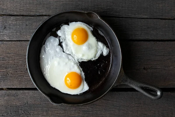 Due uova fritte in padella di ghisa isolate su vernice scura — Foto Stock