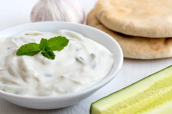 Tzatziki in white ceramic bowl with mint leaf garnish next to pi — Stock Photo, Image