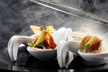 Beyaz seramik kaşıklardaki kanepeler duman içinde. Siyah arkaplan.