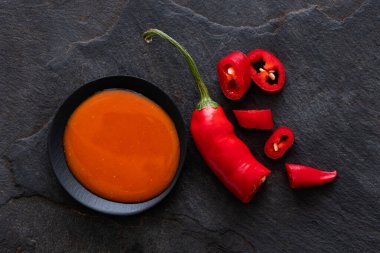 Peri peri kırmızı biber sosu siyah bir seramik kasede, dilimlenmiş bir tabağın yanında. 