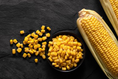 İki mısır koçanı yanında siyah bir seramik kasede konserve mısır. 