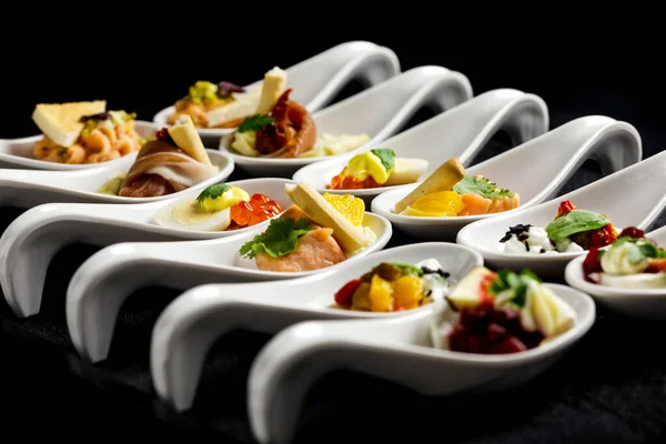 Dos filas de canapés en cucharas de cerámica blanca. Fondo negro . —  Fotos de Stock