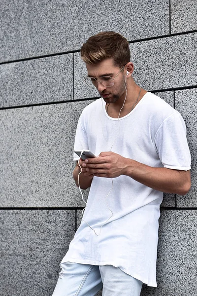 People, technology, travel and tourism - man with earphones, smartphone on city street and listening to music over gray wall background . Earphones concept.