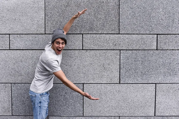 Cheerful guy dressed in gray t-shirt, sunglasses and hat at the street, show a big sale, or nice offer. Advertise concept. People, travel and tourism - man on city street show with his hands something over gray wall background