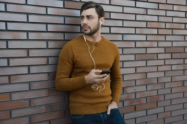 Portrait of smiling man with headphones and cellphone standing by brick wall.