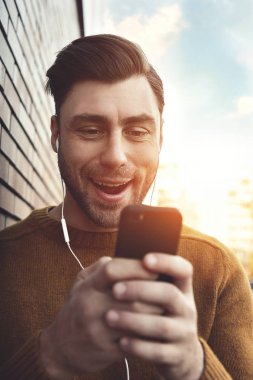 Kulaklıklar müzik dinlemek ve tuğla duvara yaslanmış gülümseyen adam. Adam cep telefonu ayakta ve kulaklık ile tuğla duvar tarafından gülümseyen portresi.