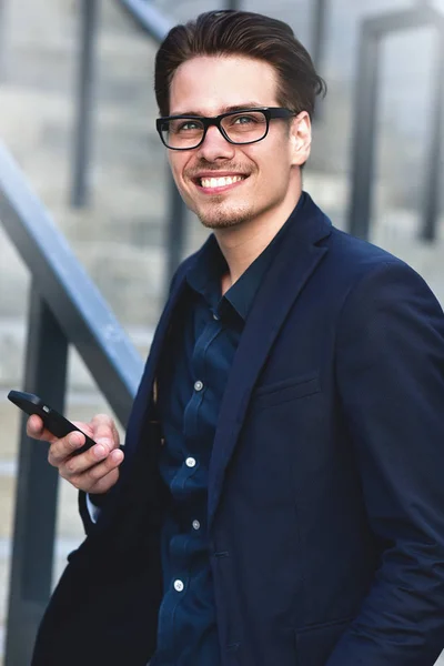 Exitoso hombre de negocios en gafas se para con el teléfono en una ciudad moderna . — Foto de Stock