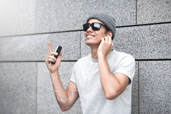 Веселый парень, одетый в серую футболку, солнцезащитные очки и шляпу на улице, слушающий музыку с наушниками, держащий мобильный телефон . — стоковое фото