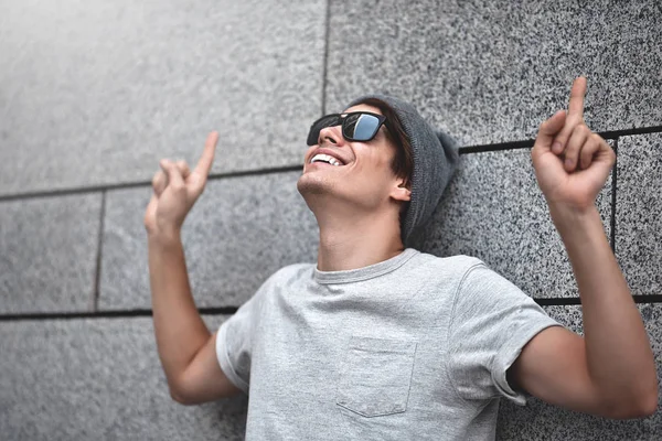 Handsome young man with sunglasses is pointing away, looking up and smiling, standing against gray wall.