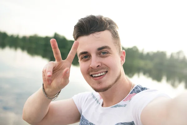 Portrait de jeune beau gars souriant élégant fait selfie contre le lac. signe de paix — Photo