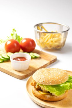 Yuvarlak ahşap tabakta kızarmış tavuk burgeri yanında patates kızartması ve beyaz arka planda ketçap servis ediliyor..