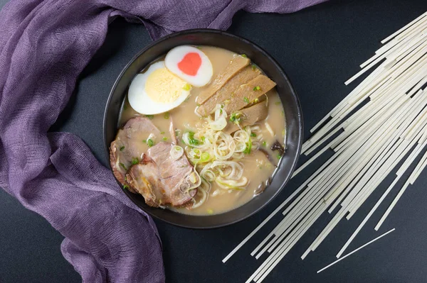 Delicious tonkotsu ramen, Japanese noodle in pork Bone based soup topped with chashu pork, boiled egg, pickled bamboo shoot and spring onions. top view on black background Decorated with purple napkin