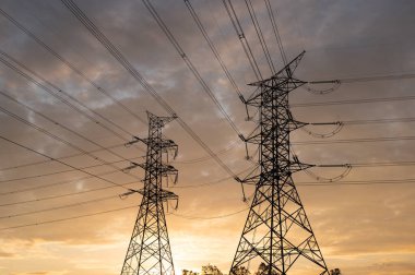 Yüksek voltajlı elektrik direklerinin ve çizgilerin silueti, günbatımı gökyüzü arka planında yüksek voltajlı elektrik iletim kulesi..