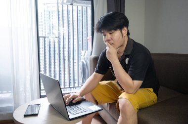 Stresli Asyalı iş adamı evdeki kanepede oturuyor ve dizüstü bilgisayarla çalışıyor. Yorgun adam ev konseptinden çalışıyor..