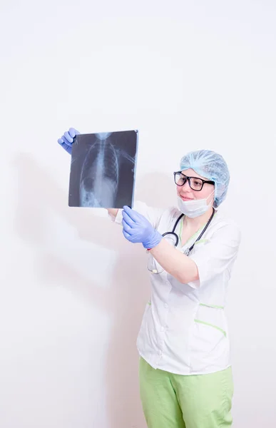 A doctor in a white coat, green trousers, protective mask, blue medical gloves, with a stethoscope around his neck, looking at a chest x-ray. on white background. — Stock Photo, Image