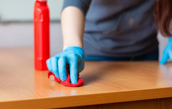 Gri kazaklı ve mavi eldivenli genç temizlikçi kadın. Ahşap bir masayı kırmızı bir bezle yıkıyor.