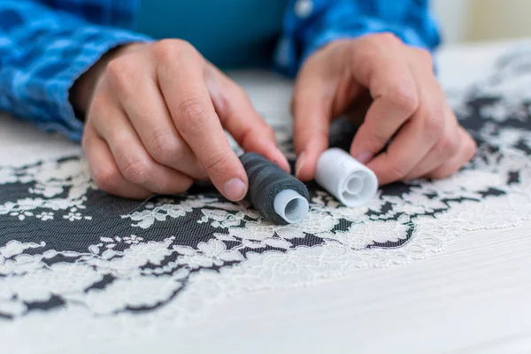 Mavi kareli gömlekli neşeli bir terzi atölyesinde çalışıyor. Yakın plan, dikiş makinesinin yanındaki masada iç çamaşırı için dantel kumaştan iplikler seçer..