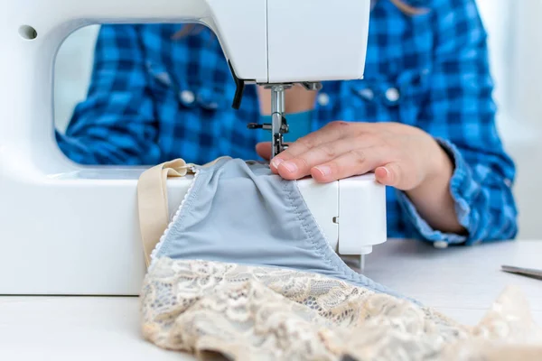 Una hermosa costurera alegre mujer en una camisa a cuadros azul, en su taller, en una máquina de coser, cosiendo un sujetador azul con inserciones beige. De cerca. —  Fotos de Stock