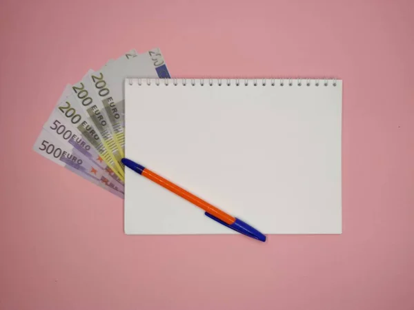 На розовом фоне, ручка по диагонали на блокноте с копировальным местом, 200 и 500 евро купюры выглядывают из-под него. горизонтальное фото . — стоковое фото