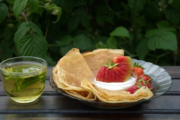 En una mesa de madera oscura hay un plato de panqueques doblados en un triángulo, y un platillo de crema con una gran fresa flotando en ella. junto a ella es transparente taza de té con grosella negra y hojas de menta . — Foto de Stock