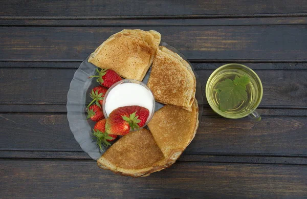 En una mesa de madera oscura hay un plato de panqueques apilados en un triángulo, y una taza de crema con una fresa grande flotando en ella. al lado hay una taza de té transparente con grosella negra y hojas de menta — Foto de Stock