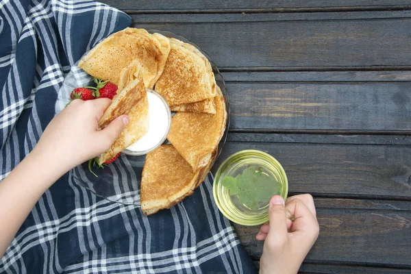 Primer plano de las manos de la persona comiendo desayuno en la mesa de madera oscura con plato de panqueques y platillo de crema y fresas.al lado de ella es transparente taza de té con grosella negra y hojas de menta . — Foto de Stock