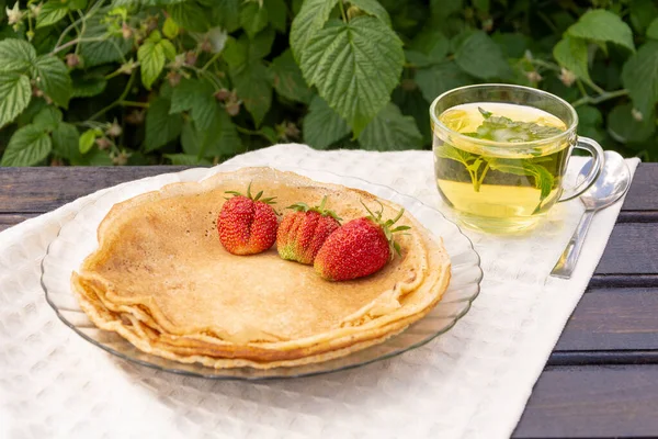 Sobre una mesa de madera cubierta con un mantel blanco, hay un plato con panqueques, y en el borde de una fresa una taza de té con grosella negra y hojas de menta. sobre un fondo de follaje verde — Foto de Stock