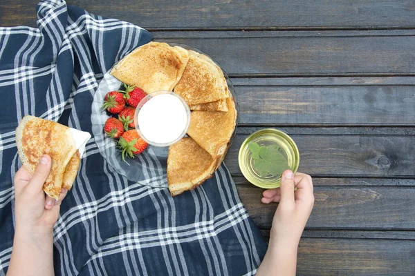 Primer plano de las manos de la persona que come el desayuno en una mesa de madera oscura con un plato de panqueques y un platillo de crema y fresas.junto a ella es una taza transparente con té verde y una hoja de menta . — Foto de Stock