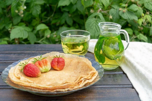 En una mesa de madera hay un plato con panqueques en el que hay varias fresas, junto a una jarra transparente y una taza con té verde y hojas de grosella negra y menta . — Foto de Stock