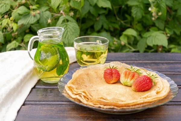 En una mesa de madera oscura hay un plato de panqueques apilados en un triángulo, y una taza de crema con una fresa grande flotando en ella. al lado hay una taza de té transparente con grosella negra y hojas de menta — Foto de Stock