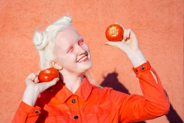 Kırmızı arka planda kırmızı bluzlu komik albino kız. Gözlerinin önünde iki kırmızı domates tutuyor ve geniş bir şekilde gülümsüyor. Boşluğu kopyala.