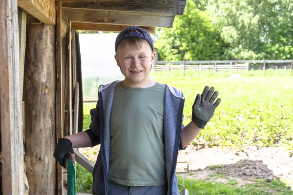 Yeşil tişörtlü, şapkalı ve iş eldivenli bir çocuk. Bahçede, çatının altında. Çocuk asistan neşeyle el sallıyor. .