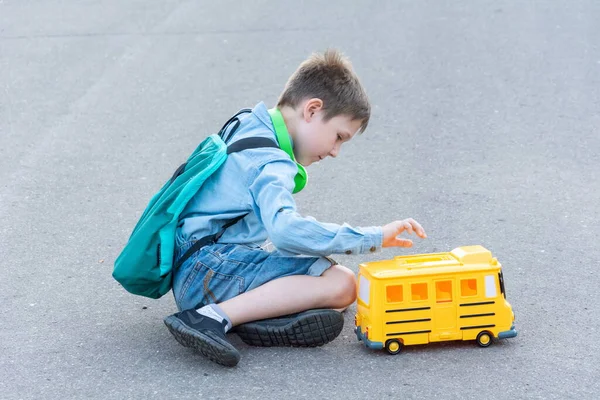 A boy 7-8 years old, sitting on the asphalt and playing with a toy school bus. Back to school concept. — Stock Photo, Image