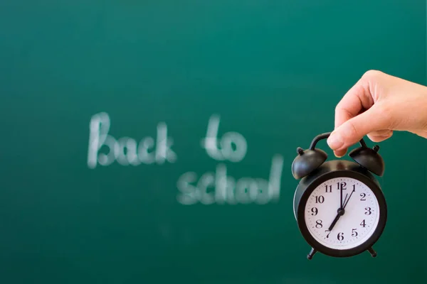 En la palma de la mano, sobre el fondo de una pizarra con la inscripción de vuelta a la escuela, enfoque suave, hay un reloj despertador negro. Regreso al concepto escolar. — Foto de Stock