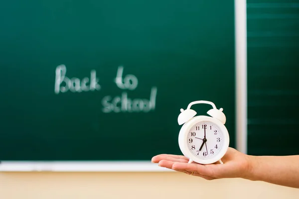 Op de palm van je hand, tegen de achtergrond van een schoolbord met de inscriptie terug naar school, staat een witte wekker. zachte focus. Terug naar schoolconcept. — Stockfoto