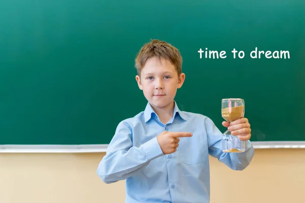 A boy in a blue shirt stands at a Board that says, time to dream. he holds an hourglass in his hand and points at it with his finger. — Stock Photo, Image