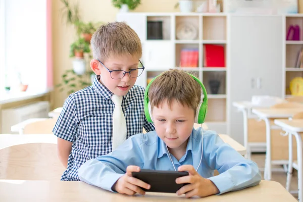 Een schooljongen in een blauw shirt speelt aan de telefoon, een andere leerling staat achter zijn schouder en kijkt toe — Stockfoto