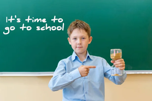 Un chico con una camisa azul se para en una pizarra que dice que es hora de ir a la escuela. sostiene un reloj de arena en su mano y lo señala con su dedo. — Foto de Stock