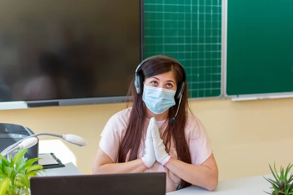 Mavi tıbbi maskeli, kulaklıklı, beyaz eldivenli, elini bir arada tutup yardım için dua eden yorgun, genç bir öğretmen. Vücut dili kavramı..