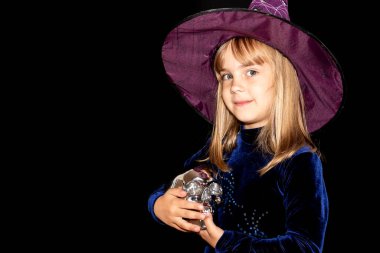  Siyah arka planda, kafasında kocaman bir şapka olan küçük cadı. Kızın elinde gümüş bir kafatası var. Cadılar Bayramı kostümü.