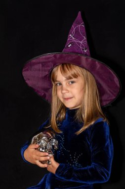  Siyah arka planda, kafasında kocaman bir şapka olan küçük cadı. Kızın elinde gümüş bir kafatası var. Cadılar Bayramı kostümü.