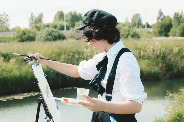 Siyah şapkalı genç bir kadın nehir kıyısında duruyor ve fırçayla tuvale bir manzara çiziyor..