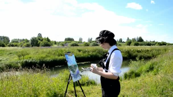 Uma jovem mulher em um boné preto fica na margem de um rio e pinta uma paisagem em tela com uma escova. — Vídeo de Stock
