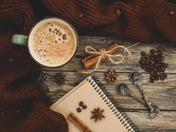 Gemütlich gestrickter brauner Pullover und aromatische Tasse Kaffee, Zimt, Anissterne, Löffel, Kaffeebohnen und Notizblock auf Holzgrund. Herbst oder Winter Compositoin. Stimmung, Lebensstil, Stilllebenskonzept. — Stockfoto